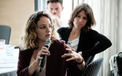 A woman with curly blonde hair and glasses speaks into a microphone during a discussion at a professional event. Two other participants, a man and a woman, listen attentively in the background.