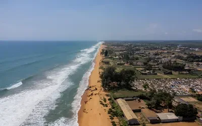 Aerial view of a long sandy coastline near the Benin–Togo border, with ocean waves along the shore on one side and a coastal settlement with scattered buildings, trees, and roads on the other.