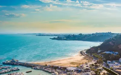 Panoramic coastal view of Sidi Bou Said near Tunis, Tunisia, showing a curved sandy beach, turquoise sea, a marina with small boats in the foreground, and white buildings along a hillside under a partly cloudy sky.