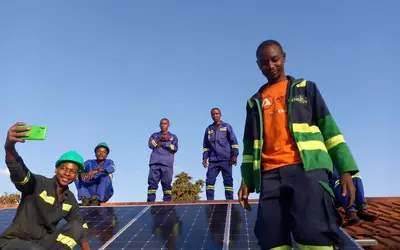 A small group of people wearing high‑visibility safety vests and hard hats stand on an elevated structure outdoors, with one person in the foreground and others behind raising their arms against a bright sky.