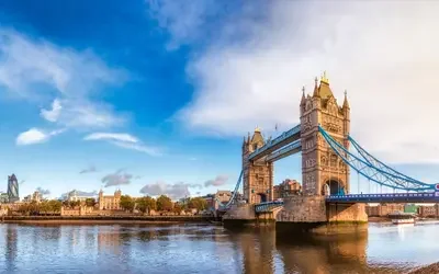 Panoramic view of London’s Tower Bridge spanning the River Thames, with blue suspension cables and two stone towers, the city skyline including the Tower of London in the background, and a partly cloudy sky reflected on the water.