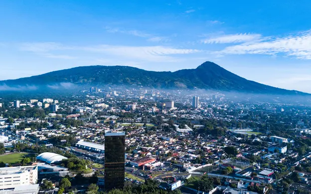 View of San Salvador, El Salvador, with urban neighborhoods and office buildings set against a mountainous landscape under a clear blue sky.