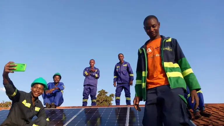 A small group of people wearing high‑visibility safety vests and hard hats stand on an elevated structure outdoors, with one person in the foreground and others behind raising their arms against a bright sky.