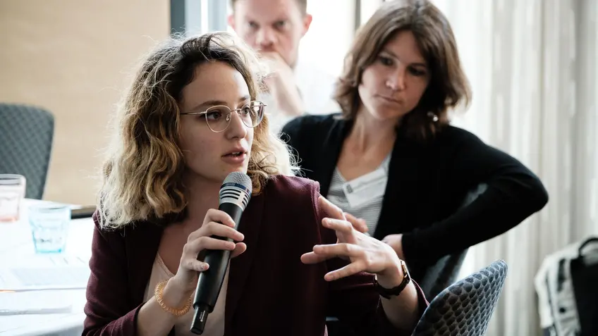 A woman with curly blonde hair and glasses speaks into a microphone during a discussion at a professional event. Two other participants, a man and a woman, listen attentively in the background.