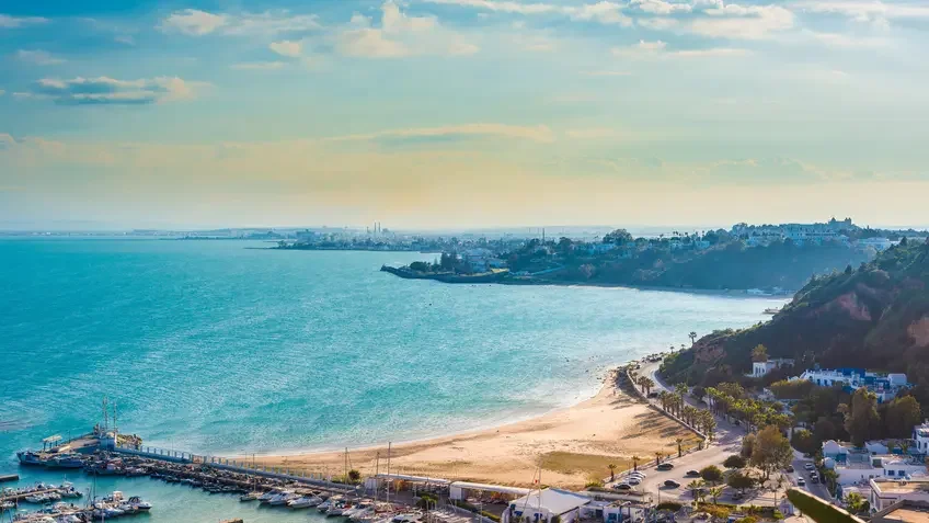 Panoramic coastal view of Sidi Bou Said near Tunis, Tunisia, showing a curved sandy beach, turquoise sea, a marina with small boats in the foreground, and white buildings along a hillside under a partly cloudy sky.