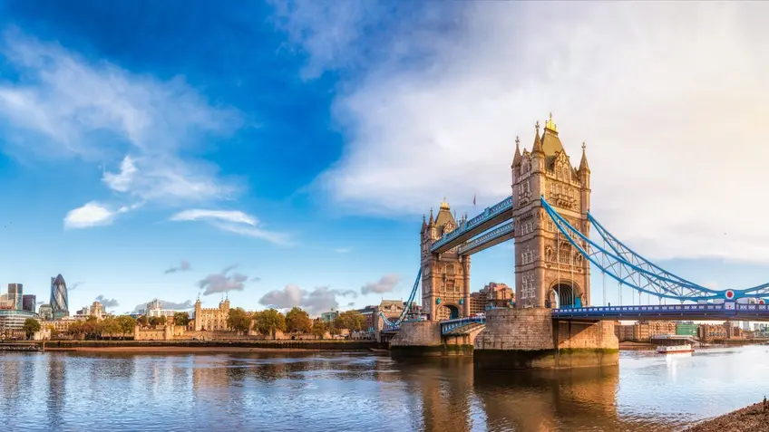 Panoramic view of London’s Tower Bridge spanning the River Thames, with blue suspension cables and two stone towers, the city skyline including the Tower of London in the background, and a partly cloudy sky reflected on the water.