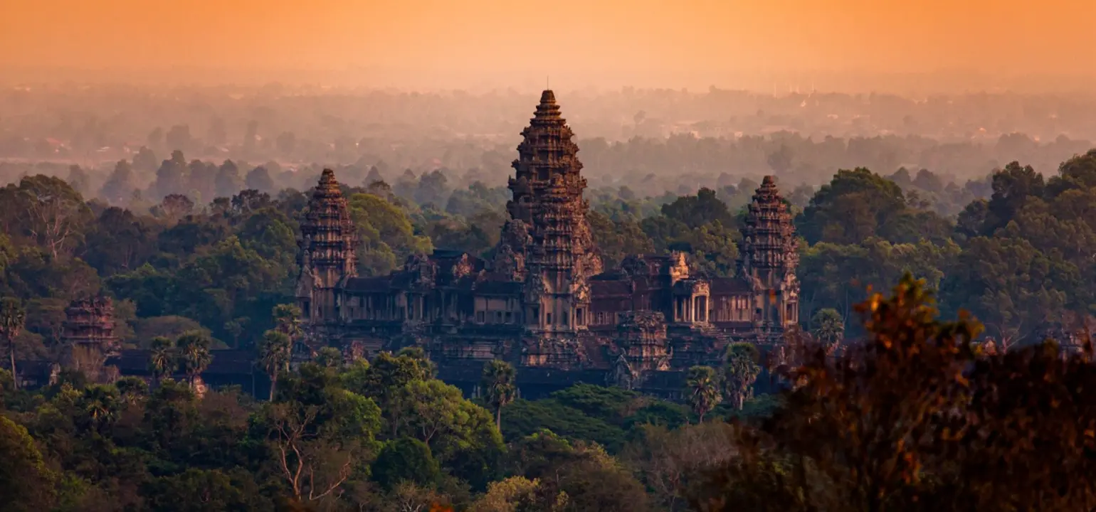 Ancient stone temple complex with tall, ornate towers rising above dense green forest, viewed from a distance at sunrise under a warm, hazy orange sky