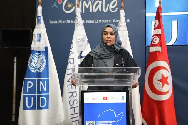 A speaker stands at a podium during the International Conference for NDC Implementation in Tunisia in 2024, with conference and national flags displayed in the background.