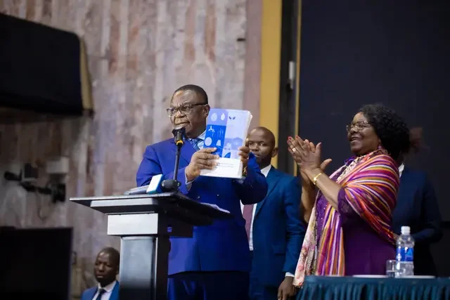 A speaker stands at a podium holding a report during a conference session, while other participants stand nearby on stage.