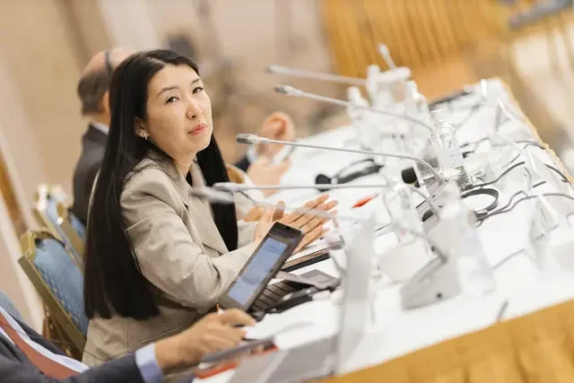 Participant speaks during a partner coordination meeting in Kazakhstan in 2025, seated at a conference table with microphones, laptops, and meeting materials.