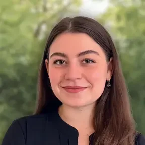 Head-and-shoulders portrait of a person with long straight brown hair parted in the middle, wearing small hoop earrings and a black top, looking directly at the camera. The background is softly blurred greenery, creating a natural, outdoor bokeh effect.